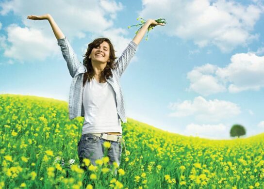 A happy, healthy looking woman in a field surrounded by yellow wild flowers.