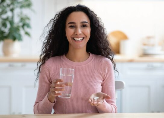 A beautiful woman smiling, getting ready to take a supplement with a glass of water