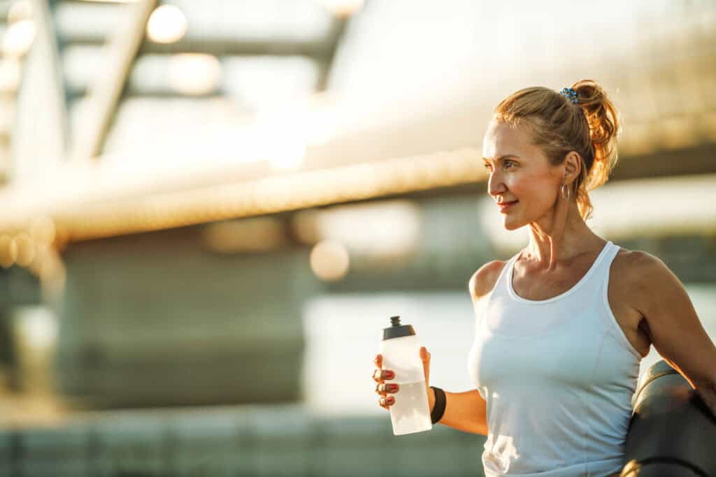 Middle age sportswoman holding water bottle before hard training near to river, in the city.
