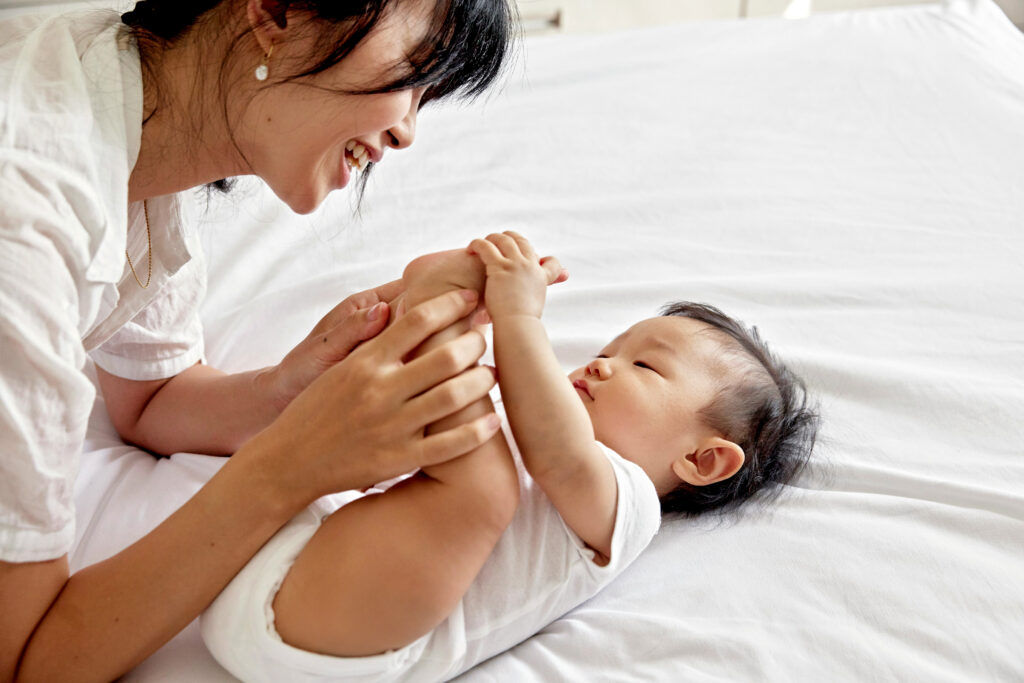 Woman smiles at baby on bed; natural light fills room.